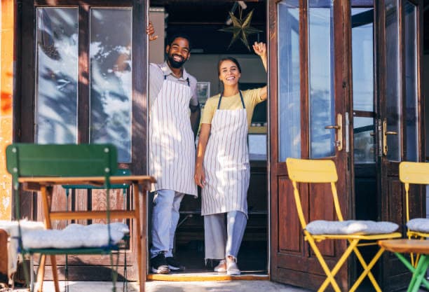 Two local shop owners in aprons waving from the open doorway of their café